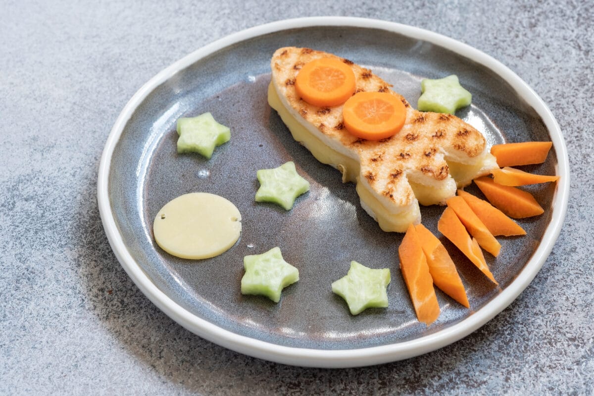 A plated sandwich shaped like a rocket with cucumber stars, a cheese moon, and carrot fire