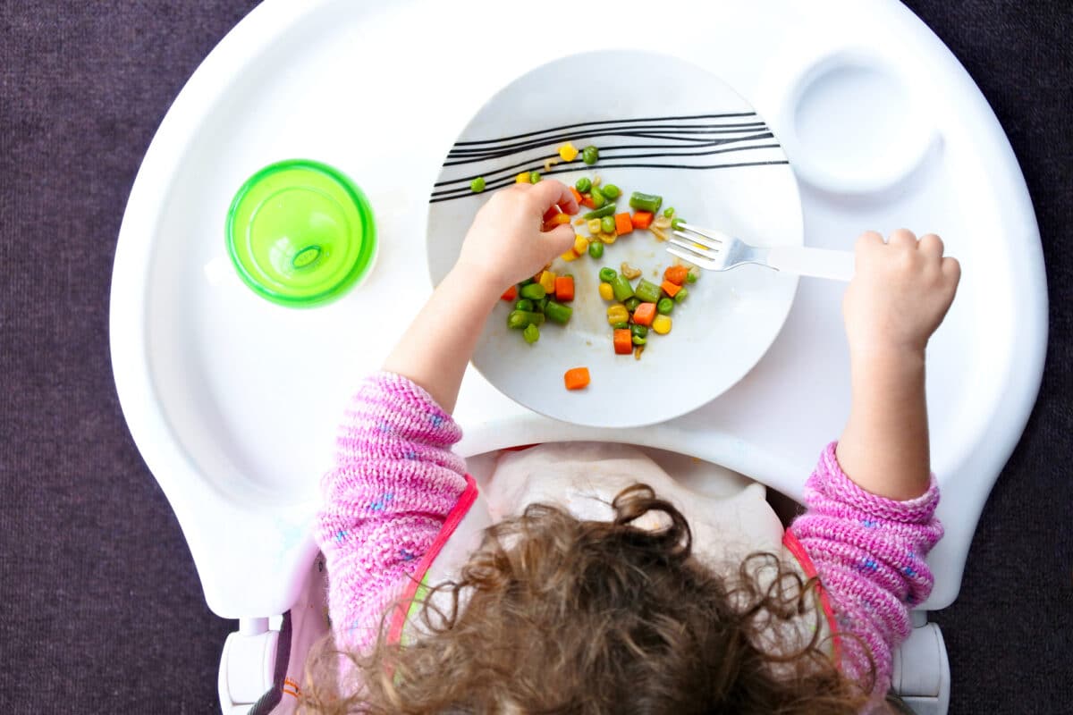 A toddler in her high chair with a plate of vegetables. She also has a green sippy cup