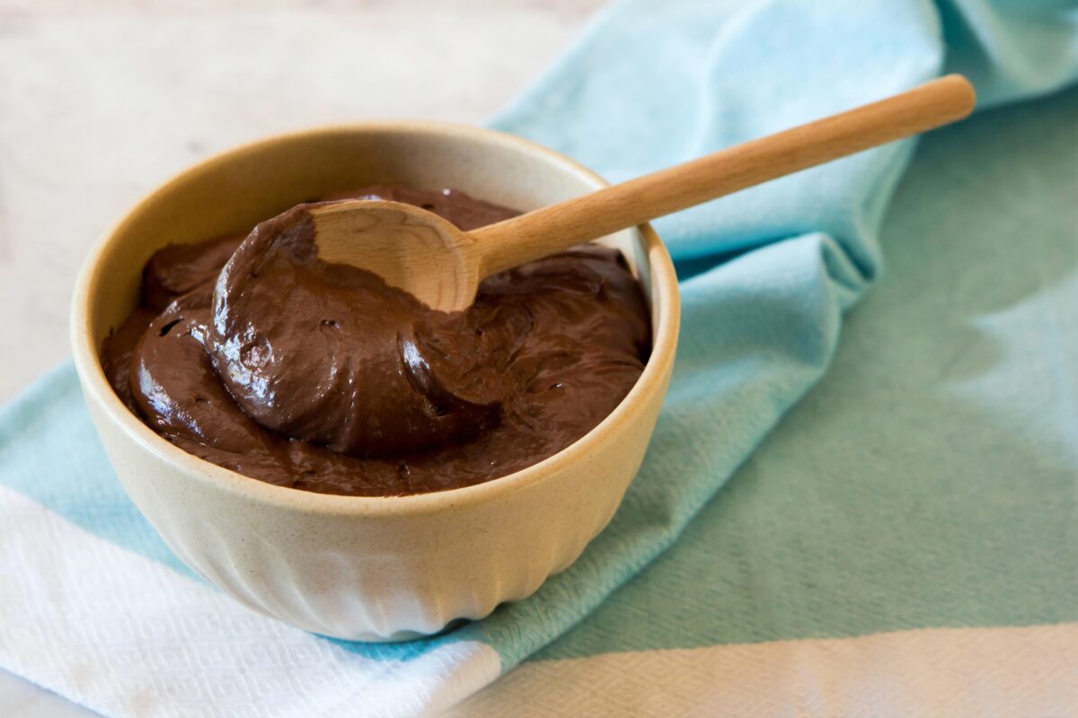 White bowl full of chocolate pudding with a bamboo spoon in the bowl. The bowl is on a blue and white tablecloth