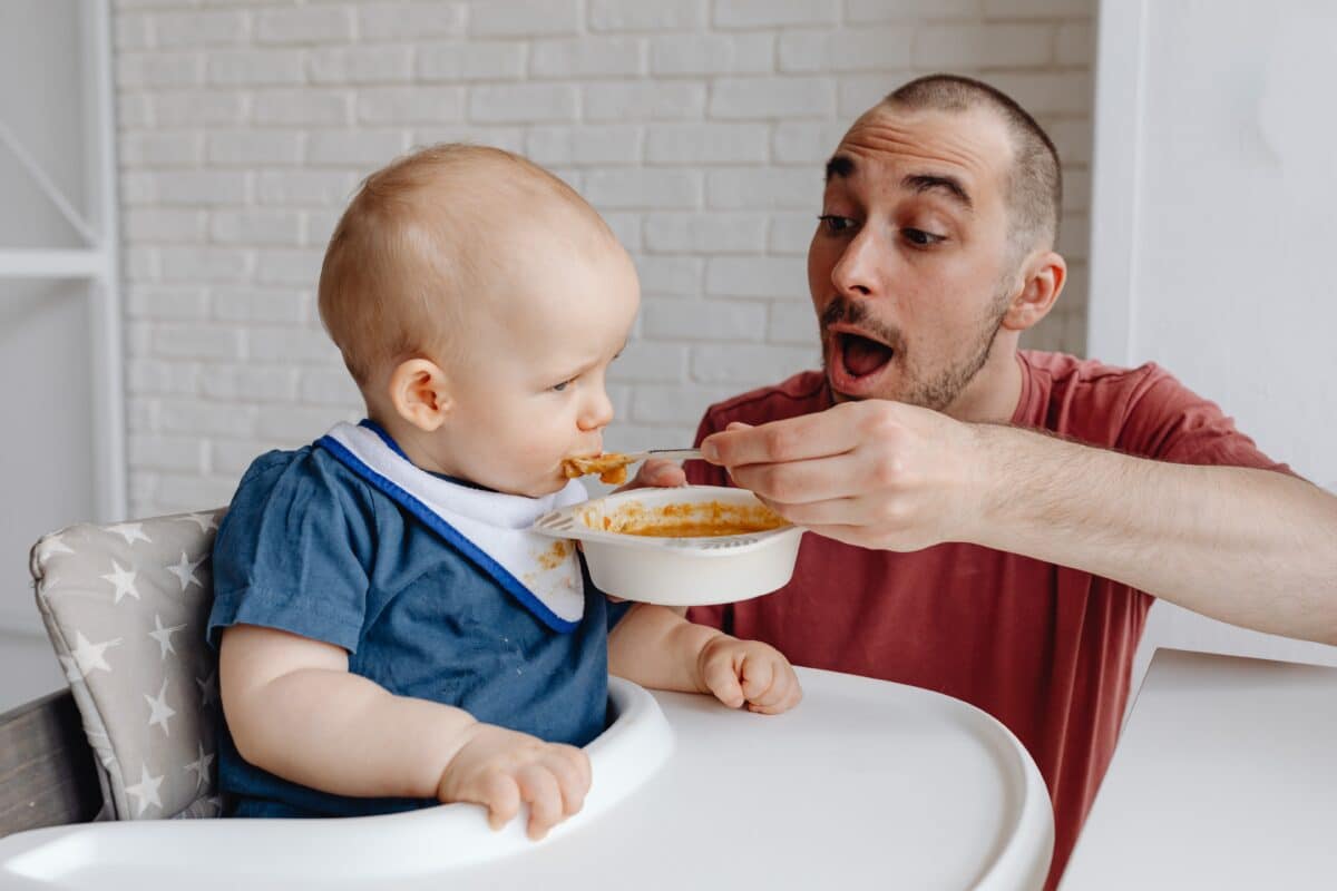 A father feeds his infant son something from a bowl. The baby is in a highchair and wearing a blue shirt and white bib.